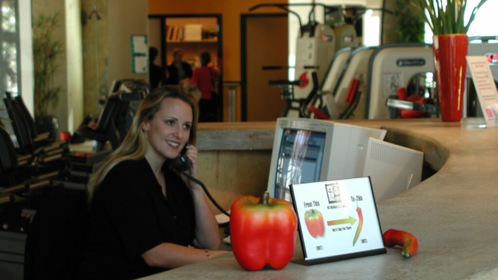 Personal trainer working at a studio front desk with exercise equipment in the background, showing the business side of a personal training studio
